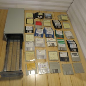 May include: A collection of vintage 3.5-inch floppy disks in various colors, with handwritten labels, alongside a gray disk storage unit. The disks are arranged on a wooden surface, showcasing a retro computing theme.