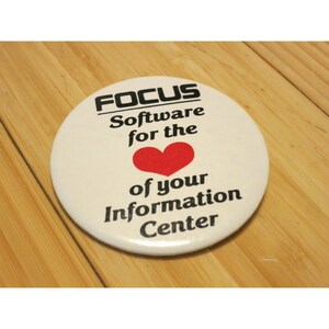 May include: A round, off-white button with the word "FOCUS" above the text "Software for the" and a red heart, followed by "of your Information Center" in black text. The button is on a light brown wooden surface.