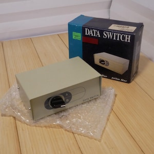 May include: A beige Data Switch device with a black knob, labeled A and B, sits on bubble wrap. A matching box with "DATA SWITCH" printed on it is behind the device. The box has a blue top and a black front with a picture of the device.