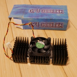 May include: A black CPU cooler with a fan and heat sink fins. The cooler has a green and black fan and is connected to a box with the words "CPU COOLER" printed on it. The cooler is designed to dissipate heat from a computer's central processing unit.