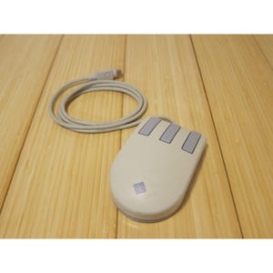 May include: A vintage computer mouse with three buttons and a cord. The mouse is off-white with light purple buttons and a matching base. The cord is coiled next to the mouse on a wooden surface.