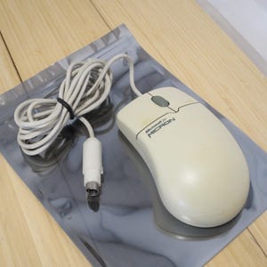 May include: An off-white computer mouse with a cord, resting on a reflective gray surface. The mouse has a gray button and the text "Microsoft for MICRON" printed on it. The cord is white and bundled with a black tie.