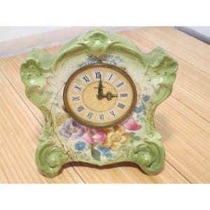 May include: A vintage mantel clock with a light green, ornate porcelain frame. The clock face is gold with black Roman numerals and hands. Floral patterns in pink, purple, and blue decorate the base.