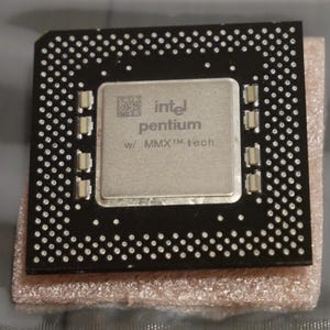 May include: A close-up of a computer processor with the Intel Pentium logo and "w/ MMX tech" text. The processor is square, with a silver center and a black border dotted with small silver connectors. It rests on a light brown surface.