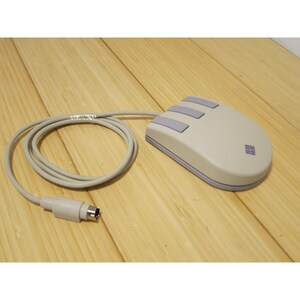 May include: A vintage computer mouse with a light gray body and three gray buttons. The mouse has a rounded shape and a gray cable. A small logo is visible on the side. The mouse is on a wooden surface.
