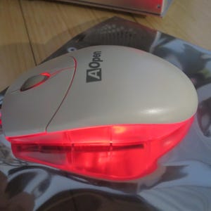 May include: A computer mouse with a cream-colored top and a red, glowing base. The mouse has a scroll wheel and the "AOpen" logo. The mouse is on a reflective surface.