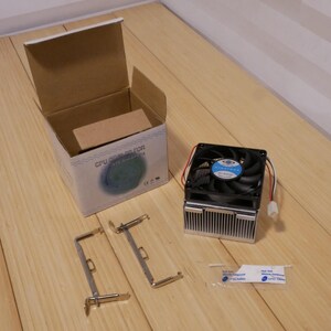May include: A computer CPU cooler kit with a black fan, silver heat sink, mounting brackets, and a box. The box is white with blue and green graphics and the text "CPU COOLER FOR". The kit is on a wooden surface.