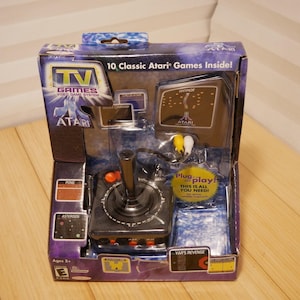 May include: A boxed Atari TV Games video game system. The box features the Atari logo, game screenshots, and the text "10 Classic Atari Games Inside!" A black joystick and game cartridges are visible inside the packaging. The box is blue and black.