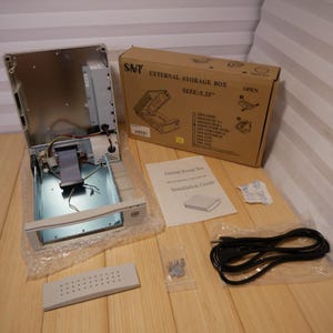 May include: An open SNT external storage box with a 5.25" size, along with its components, including a beige faceplate, screws, and a power cable. A cardboard box and an installation guide are also present.