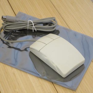 May include: A vintage off-white computer mouse with three buttons, connected to a gray cable. The mouse rests on a reflective, blue-gray mousepad. The cable is secured with a white twist tie. The background is a light-colored wooden surface.