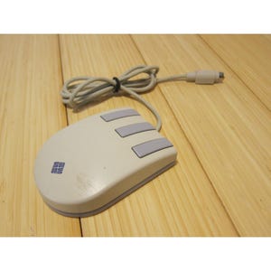 May include: A vintage computer mouse with a light gray body and three gray buttons. The mouse has a cord and a connector. The logo "SUN" is printed on the mouse. The mouse is on a wooden surface.