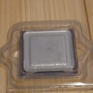 May include: A silver AMD processor in a clear plastic protective case. The processor has the AMD logo and other text visible on its surface. The case is rectangular with rounded corners, sitting on a wooden surface.
