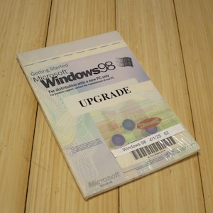May include: Sealed Microsoft Windows 98 Upgrade software package. The package is light blue and white, with the Windows 98 logo and the word "UPGRADE" prominently displayed. Includes a Microsoft sticker and a barcode.
