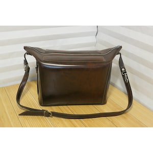 May include: A brown leather messenger bag with a rectangular shape and a long adjustable strap. The bag has a zipper closure and silver-colored hardware. The leather has a glossy finish.