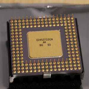 May include: Close-up of a square computer processor with gold pins. The processor has a gold-colored square center with the text "C2452733CA A4 BG 03". The processor is set on a dark gray surface.