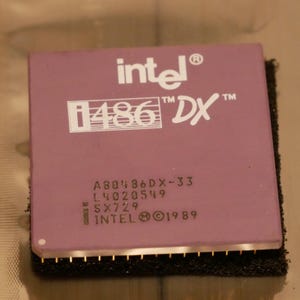 May include: A close-up of a vintage Intel i486 DX processor. The processor has a mauve-colored top with white text, including the Intel logo and the model number. The bottom edge has gold pins. The processor is set on a black foam base.