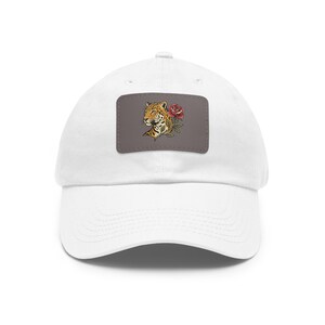 Peut inclure: Une casquette de baseball blanche avec un écusson rectangulaire sur le devant. L'écusson est gris foncé et présente une illustration détaillée d'une tête de léopard placée à côté d'une rose rouge avec des feuilles vertes et un contour noir.