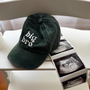 Puede incluir: Una gorra de béisbol verde oscuro con el texto bordado "big bro" en blanco. Junto a la gorra hay varias imágenes de ultrasonido, que muestran a un bebé. La gorra está sobre una superficie blanca.