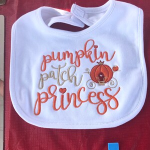 May include: White baby bib with orange embroidery that says "pumpkin patch princess" with a small pumpkin carriage in the middle.