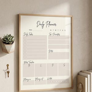May include: A framed daily planner with the words "Daily Planner" in elegant script. The planner includes sections for daily tasks, top priorities, notes, meal tracking, and water intake. The frame is light wood.