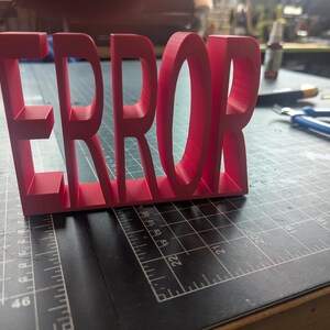 K&ouml;nnte beinhalten: Ein leuchtend rotes 3D-gedrucktes Schild mit dem Wort "ERROR". Die Buchstaben sind dick und stehen auf einer dunklen Oberfl&auml;che mit Rasterlinien. Das Schild ist der Mittelpunkt, mit Werkzeugen und anderen Gegenst&auml;nden unscharf im Hintergrund.