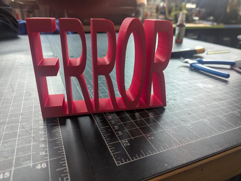 K&ouml;nnte beinhalten: Ein leuchtend rotes 3D-gedrucktes Schild mit dem Wort "ERROR". Die Buchstaben sind dick und stehen auf einer dunklen Oberfl&auml;che mit Rasterlinien. Das Schild ist der Mittelpunkt, mit Werkzeugen und anderen Gegenst&auml;nden unscharf im Hintergrund.