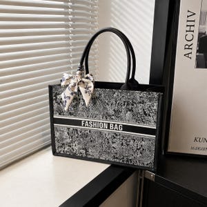 May include: A black and white rectangular tote bag featuring an intricate floral pattern, two black top handles, and a central horizontal band with the text FASHION BAG. A matching patterned ribbon is tied to one of the handles as an accessory.