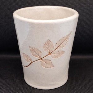 Peut inclure: Gobelet en céramique blanc cassé avec un motif de feuilles. Les feuilles et la tige sont de couleur marron clair, et le gobelet a une forme légèrement conique. Le gobelet mesure environ 13 cm de haut.