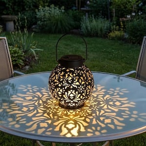 May include: A black, solar-powered lantern with a decorative floral pattern sits on a glass table. The lantern casts a patterned light onto the table's surface. The lantern has a handle for carrying and is placed outdoors.