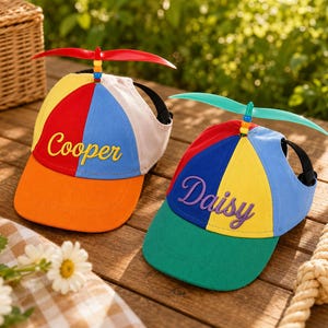 May include: Two multi-colored propeller caps featuring a red, blue, yellow, and orange panel design. The left cap has the name Cooper embroidered in yellow, and the right cap has the name Daisy embroidered in purple, both topped with a plastic propeller.