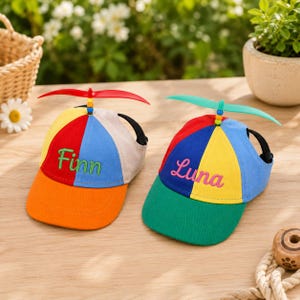 May include: Two multi-colored baseball caps with propeller attachments on top. The left cap features red, blue, yellow, and orange panels with the embroidered name Finn. The right cap has red, blue, yellow, and green panels with the name Luna.