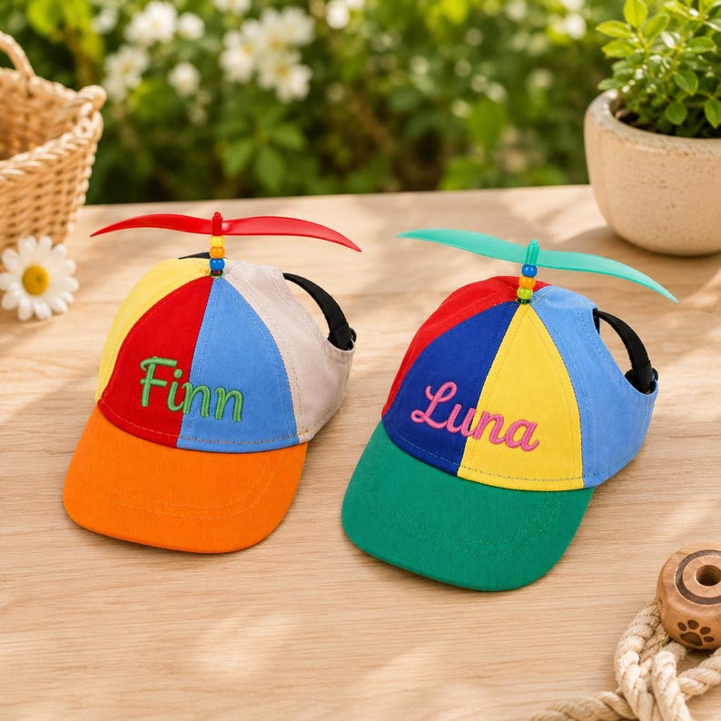 May include: Two multi-colored baseball caps with propeller attachments on top. The left cap features red, blue, yellow, and orange panels with the embroidered name Finn. The right cap has red, blue, yellow, and green panels with the name Luna.