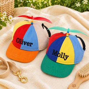 May include: Two colorful pet baseball caps with propeller tops. One cap is orange, red, yellow, blue, and tan, with "Oliver" embroidered. The other is red, blue, yellow, and green, with "Molly" embroidered. Both have black chin straps.