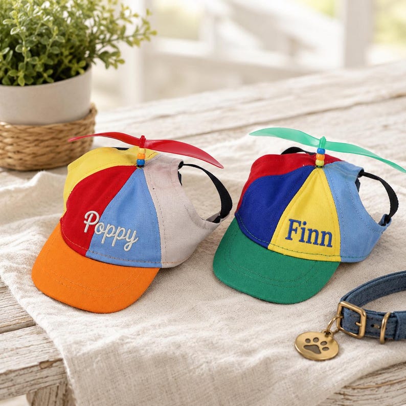 May include: Two colorful dog hats with propeller tops. One hat is orange, yellow, red, and blue with the name "Poppy". The other hat is green, yellow, red, and blue with the name "Finn". A denim dog collar with a paw print tag is also visible.