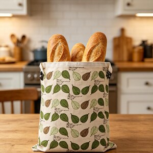 May include: A rectangular fabric bread bag featuring a repeating pattern of green and brown leaves on a cream background, with small black text interspersed between the leaves. Three golden-brown baguette loaves are tucked inside the open top.
