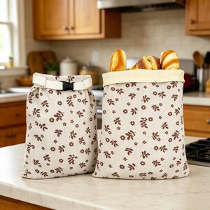 May include: Two beige fabric bags featuring a brown floral and leaf pattern. One bag is closed with a white strap and black plastic buckle, while the other is open and filled with three pieces of golden-brown bread loaves.