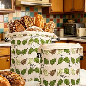 May include: Two off-white fabric bread bags featuring a repeating pattern of green and brown leaves and small black script text. The larger bag is filled with various loaves of crusty bread, while the smaller bag sits empty beside it.