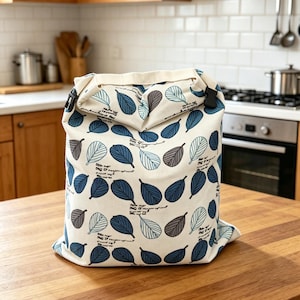 May include: A cream-colored fabric bag with a roll-top closure secured by black plastic buckles. The bag features a repeating pattern of stylized leaves in shades of dark blue, light blue, and grey, interspersed with small lines of black cursive text.