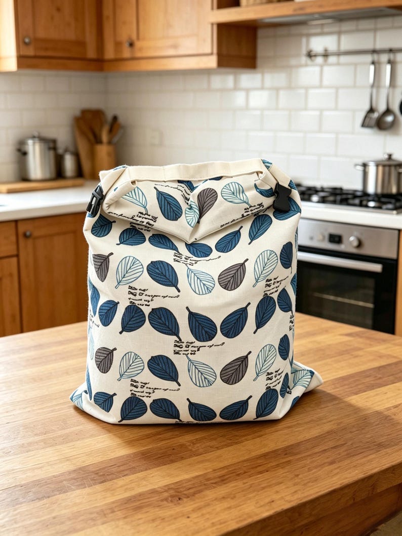 May include: A cream-colored fabric bag with a roll-top closure secured by black plastic buckles. The bag features a repeating pattern of stylized leaves in shades of dark blue, light blue, and grey, interspersed with small lines of black cursive text.