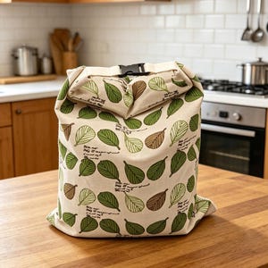 May include: A cream-colored fabric roll-top bag featuring a repeating pattern of green and brown leaves interspersed with small sections of cursive script text. The bag is secured at the top with a black plastic side-release buckle closure.