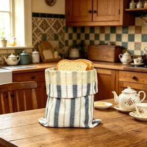 May include: A fabric bread bag featuring a vertical striped pattern in shades of blue, beige, and white. The bag has a folded top edge and contains two slices of toasted bread. The textile material has a visible weave and a structured cylindrical shape.