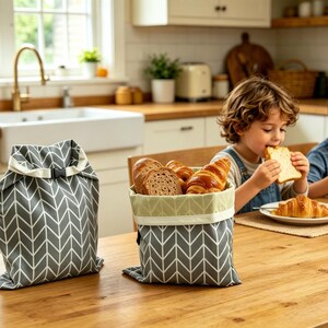 May include: Two grey and white chevron patterned fabric bags with roll-top closures and black plastic buckles. One bag is standing upright and closed, while the other is open and filled with various types of bread, including sliced loaves and croissants.