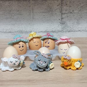 Może przedstawiać: Asortyment zdobionych jajek i szydełkowych uchwytów na jajka. Jajka są pomalowane w twarze i noszą szydełkowe kapelusze. Uchwyty na jajka obejmują jagnię, królika i pisklę. Kolory to biały, szary i żółty.