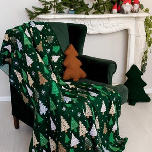 Könnte beinhalten: Eine dunkelgrüne Wohndecke mit einem sich wiederholenden Muster aus stilisierten Weihnachtsbäumen in Hellgrün, Weiß und Beige, akzentuiert durch kleine weiße Sterne. Die Decke liegt über einem dunkelgrünen Samtsessel.