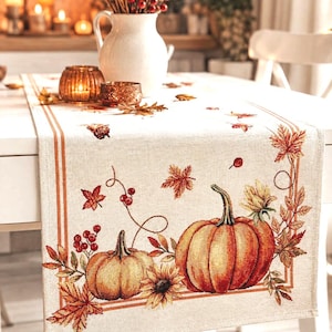 May include: A rectangular table runner in a light beige fabric featuring a woven design of two orange pumpkins, sunflowers, red berries, and scattered autumn leaves. The pattern is framed by a thin orange border on a textured linen-like material.