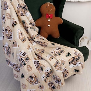 Pode incluir: Uma manta de lã bege com um padrão repetitivo de casas de gengibre, flocos de neve brancos e pinheiros verdes, sobre uma poltrona verde-escura com um peluche de boneco de gengibre tricotado com uma gravata borboleta vermelha.