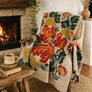 Op de afbeelding: Een beige plaid gedrapeerd over een houten fauteuil, met een herhalend patroon van grote oranje bloemen, gele chrysantachtige bloemen en donkergroen blad. Het textiel heeft een geweven structuur en bedekt de zitting en de rugleuning.