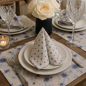 May include: A table setting featuring beige placemats with a blue floral pattern, white ceramic plates, and a beige napkin with blue polka dots folded into a triangle. The setting includes crystal wine glasses, a silver fork, and a black vase.