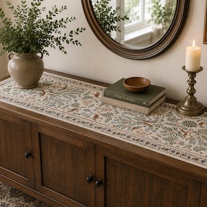 May include: A rectangular table runner featuring a detailed botanical print with green leaves, brown branches, and small birds on a cream background, bordered by a dark floral pattern, displayed on a dark brown wooden cabinet with three doors.