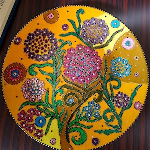 Puede incluir: Un plato decorativo pintado a mano con un diseño floral vibrante. El plato presenta un fondo amarillo con flores coloridas en tonos de rosa, morado y azul. Los tallos y hojas verdes conectan las flores. El plato es redondo.
